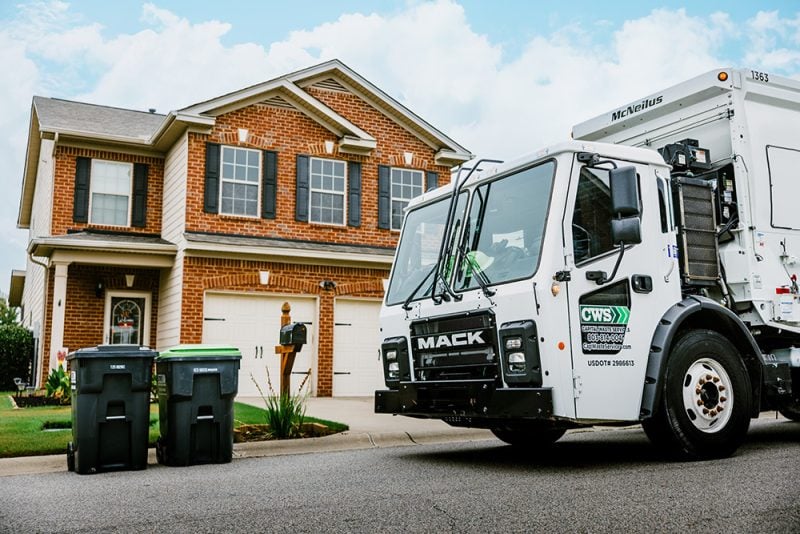 Collection truck driving by a brick house with the trash and recycle bins by the street